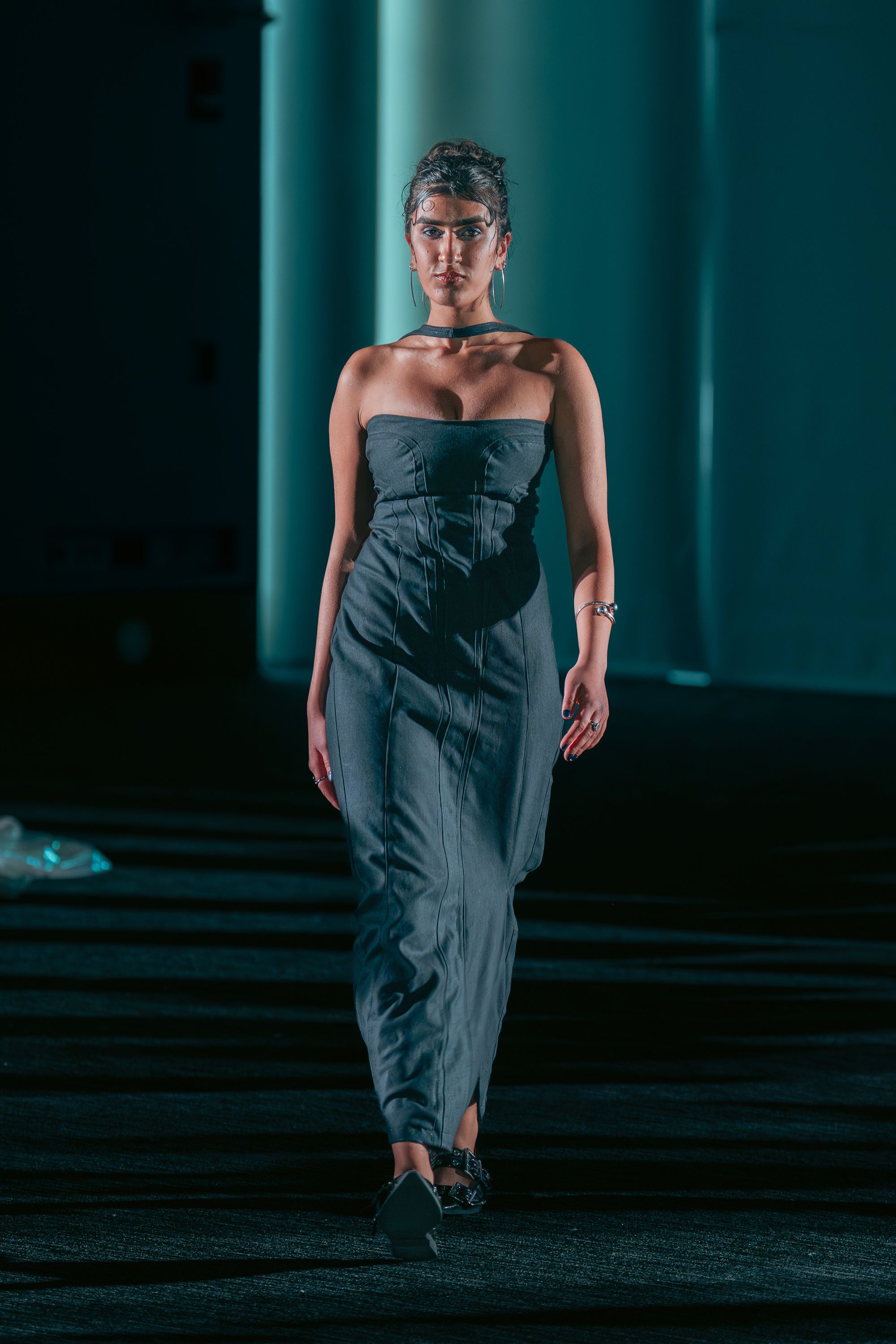 Model walks the runway in a gray strapless gown with structured vertical seam lines and a choker connected to the top of the dress. She accessorizes with hoop earrings, a silver bracelet, and strappy black heels.